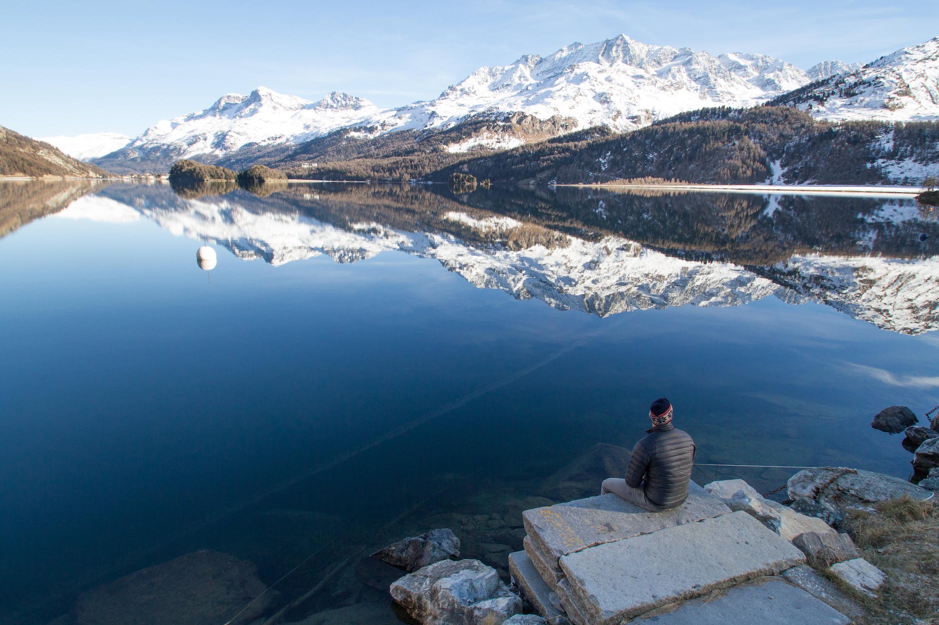 man sitting at the edge of rock facing the lake during day